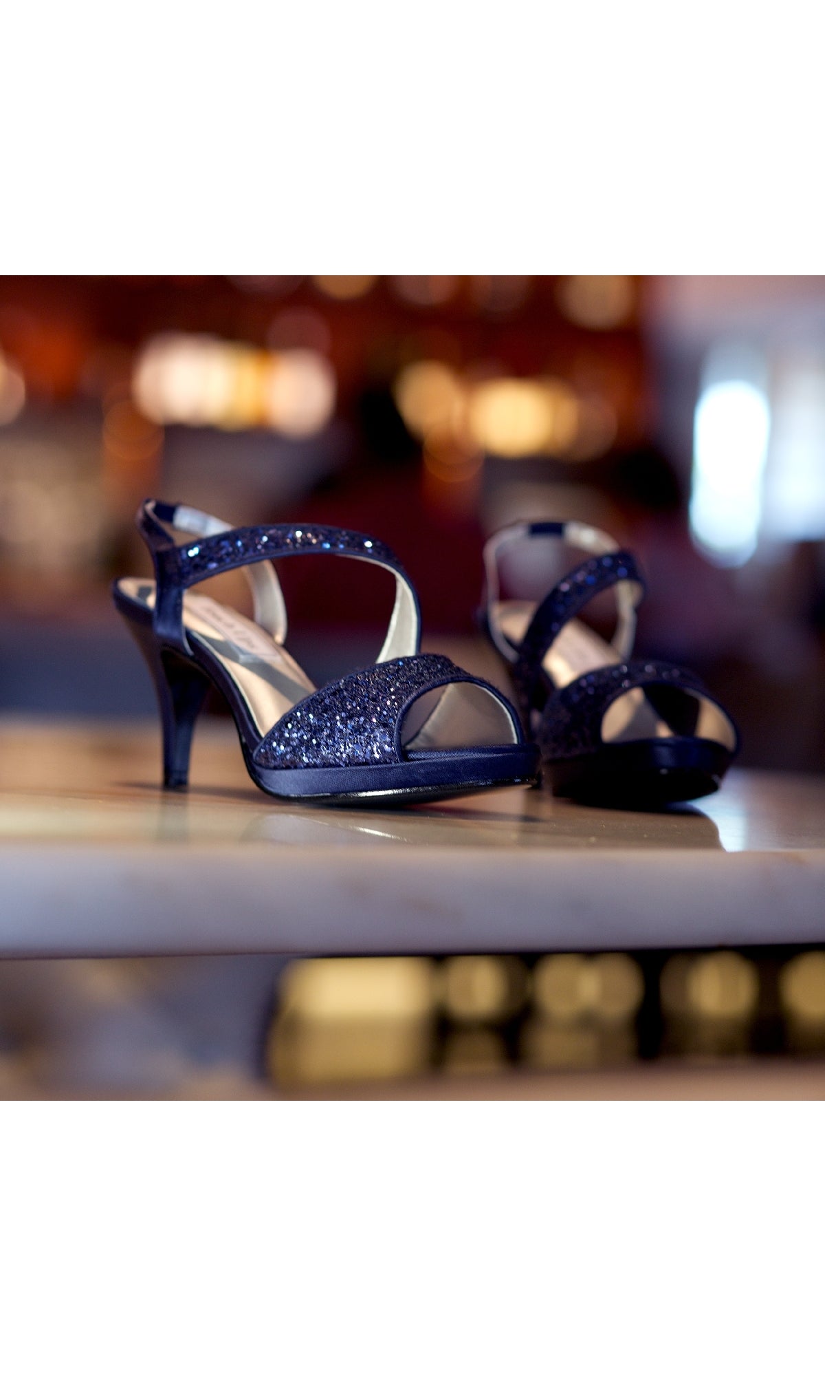 The Reagan Navy Prom Shoes (Touch Ups 4125) are chic, glittery, open-toe high-heeled sandals in navy blue, beautifully displayed on a reflective indoor surface.